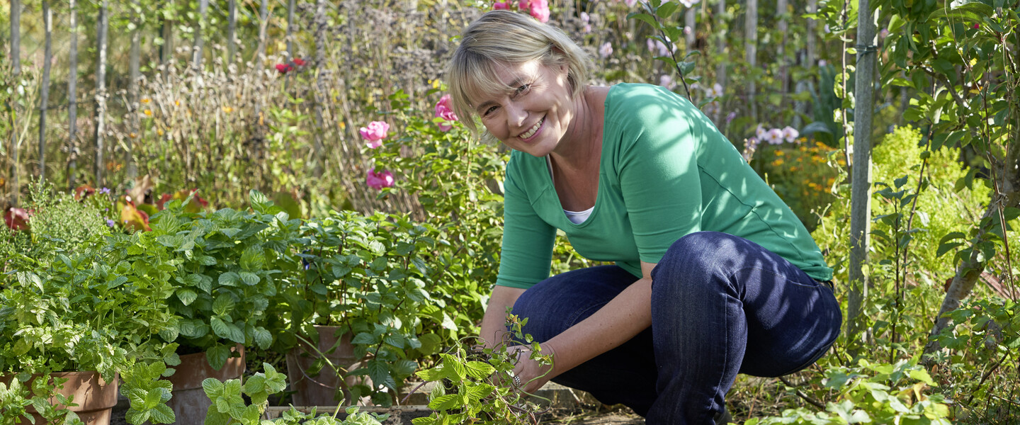 Frau hockt neben Minzekasten im Beet und schaut in die Kamera