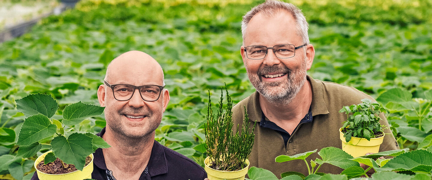 Zwei M&auml;nner halten Kr&auml;uter in T&ouml;pfen in den H&auml;nden und l&auml;cheln