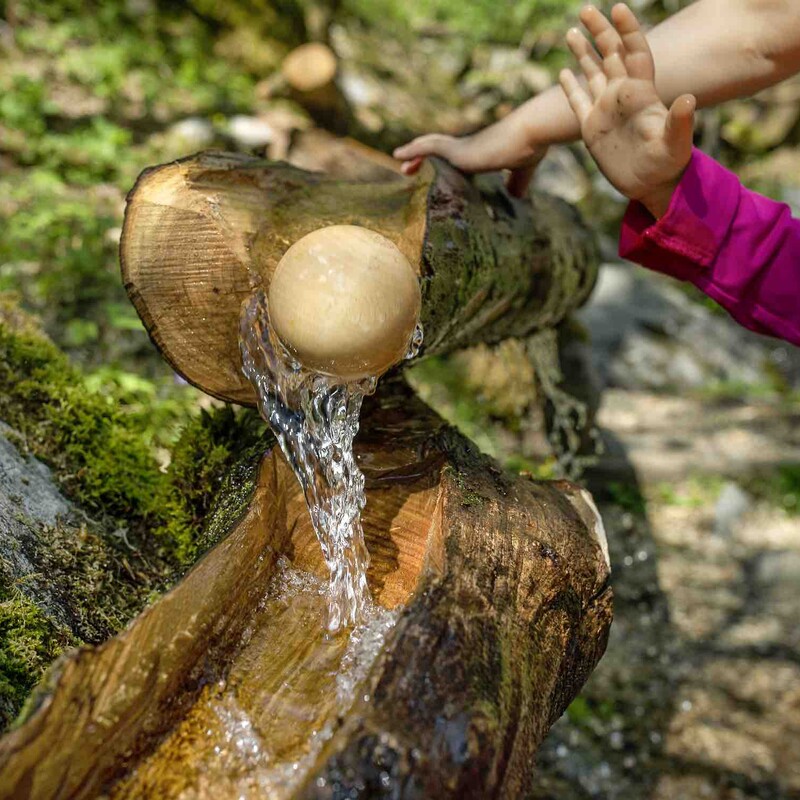 Kinderhände an einem Holzstamm, durch den Wasser fließt und eine Holzkugel rollt