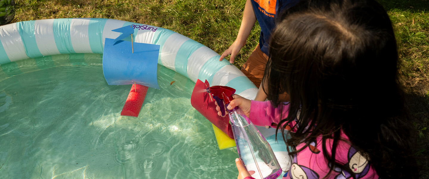 Zwei Kinder spielen mit Sprühflasche und selbstgebastelten Booten aus Poolnudeln in einem Kinderpool