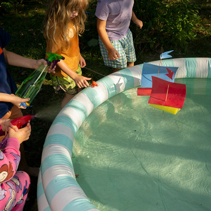 Kinder spielen mit selbsgebastelten Booten aus Poolnudeln im Planschbecken im Garten