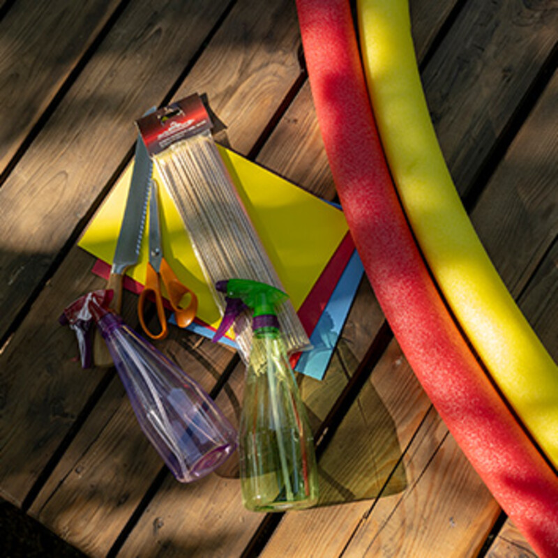 Material für Boote aus Poolnudeln: Moosgummi, Schaschlikspieße, Sprühflaschen, Schere und Brotmesser liegen auf einer Holzterrasse