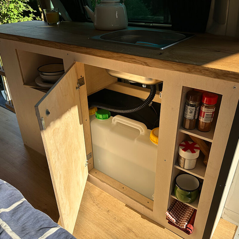 Selbstgebaute Camper-Küche mit Spüle und geöffneter Tür und einem Wasserkanister