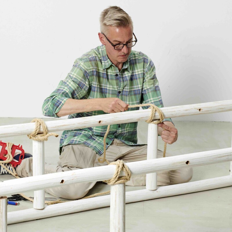 Mann sitzt auf Boden und verbindet dünne Holzstämme zu einer Daybed-Konstruktion