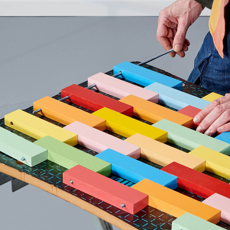 Mann schiebt eine Gewindestange durch bunt lackierte Holzleisten, die eine DIY Fußmatte bilden