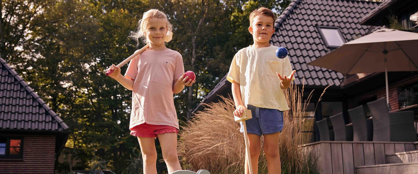 zwei Kinder stehen im Garten und halten Krocket-Schl&auml;ger und bunte Kugeln in den H&auml;nden