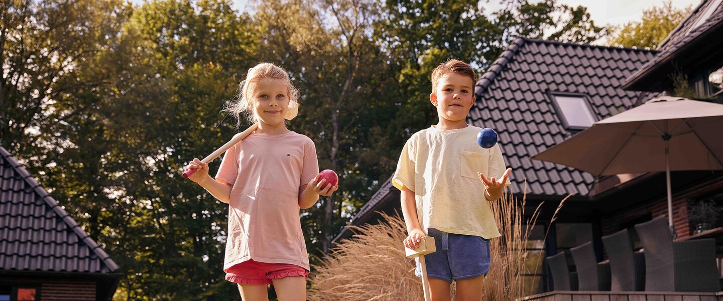 zwei Kinder stehen im Garten und halten Krocket-Schläger und bunte Kugeln in den Händen