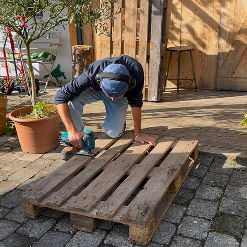 Fahrradständer aus Palette selber bauen: Mann zersägt mit Stichsäge eine Palette auf der Oberseite