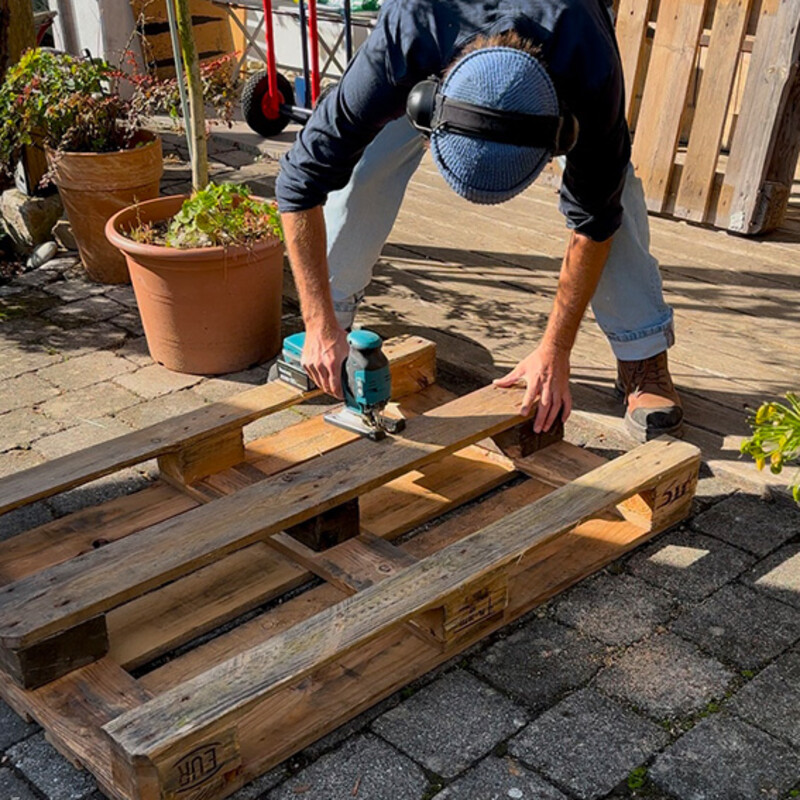 Fahrradständer aus Palette selber bauen: Mann zersägt mit Stichsäge eine Palette in der Mitte