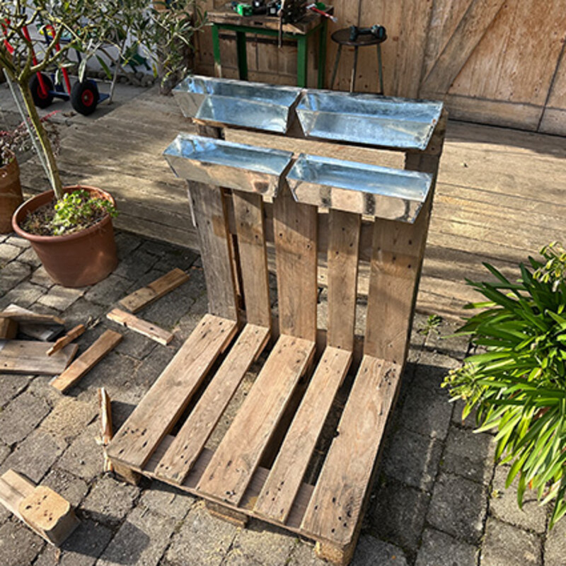 Fahrradständer aus Paletten selber bauen: Unbepflanzte Blumenkästen aus Metall hängen an senkrecht stehender Palette