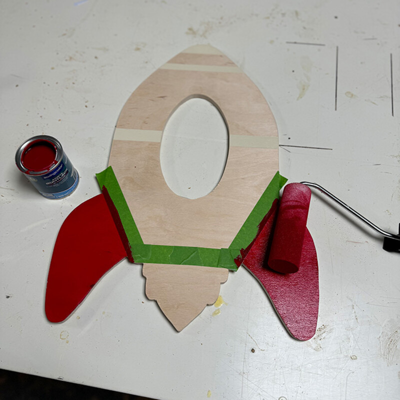 Farbrolle mit roter Farbe liegt auf Flügel von DIY-Rakete aus Holz