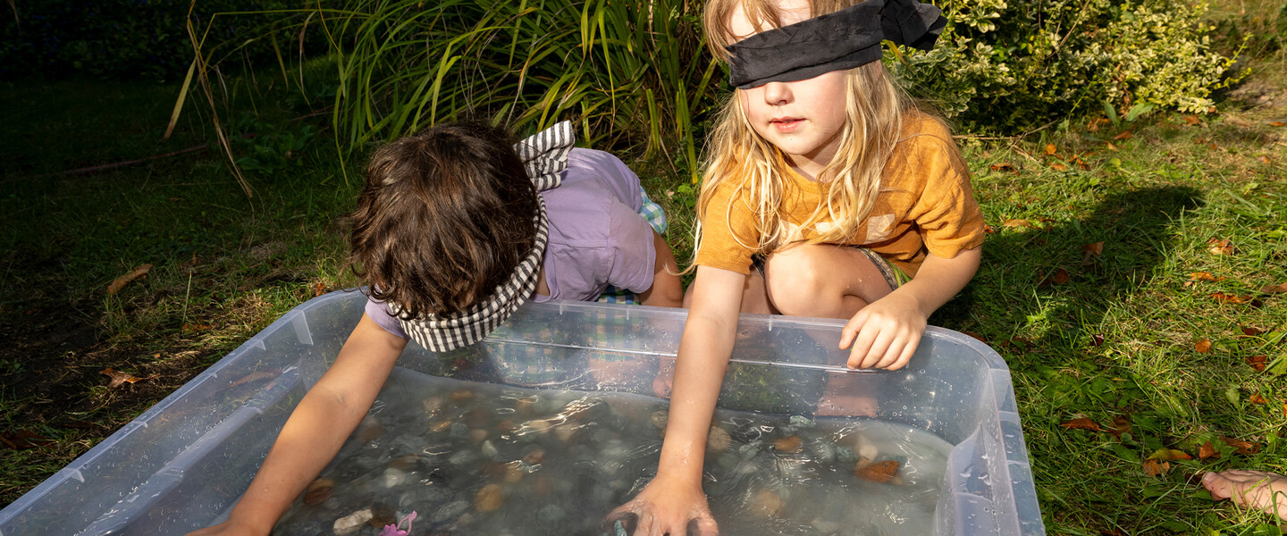 Zwei Kinder tasten mit Augenbinden in einer großen Plastikwanne mit Wasser und Steinen
