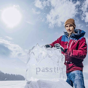Mann in roter Jacker präsentiert Eisskulptur in Form von Berggipfeln auf einem zugefrorenen See