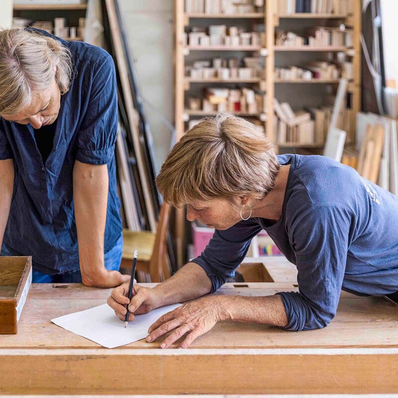 Zwei Frauen beugen sich über Werkbank und zeichnen etwas auf Papier
