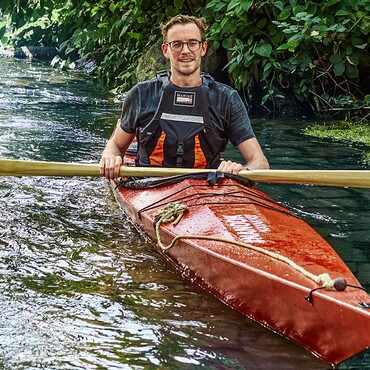 Mann mit Brille sitzt im roten Kanu auf dem Wasser und hält ein Paddel in der Hand