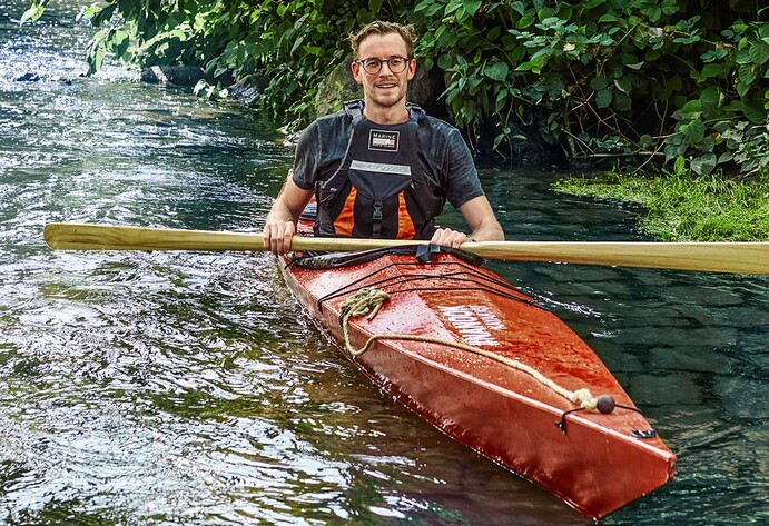Mann mit Brille sitzt im roten Kanu auf dem Wasser und hält ein Paddel in der Hand