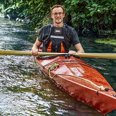 Mann mit Brille sitzt im roten Kanu auf dem Wasser und hält ein Paddel in der Hand