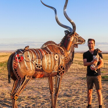 Mann mit Hund steht neben Metall-Skulptur einer Antilope