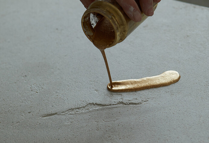 Hand hält Glas und gießt daraus goldene Farbe auf eine Platte