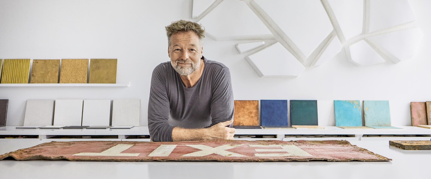 Uwe Schmidt stützt sich auf Tisch vor einer Wand mit bunten Holzplatten