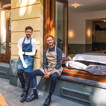 Zwei Männer mit Restaurantschürzen vor Restaurant mit offenem Fenster
