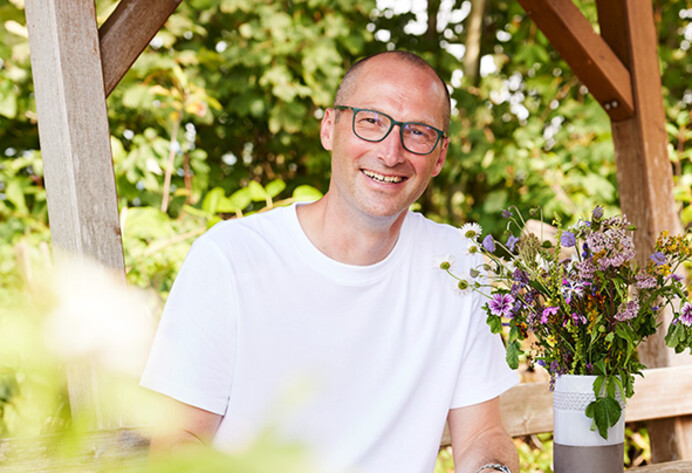 Mann mit weißem T-Shirt und Brille (Johannes Huber von der der Firma Samen Maier) sitzt im Garten an einem Holztisch.