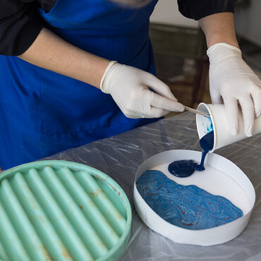 Frau mit Plastik-Handschuhen gießt blaue Farbe auf einem Becher in eine Schale