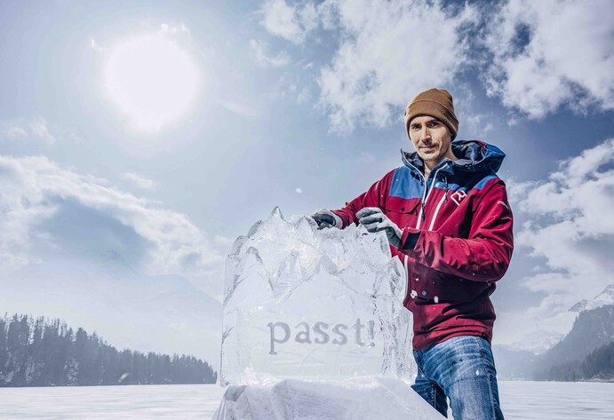 Mann in roter Jacker präsentiert Eisskulptur in Form von Berggipfeln auf einem zugefrorenen See