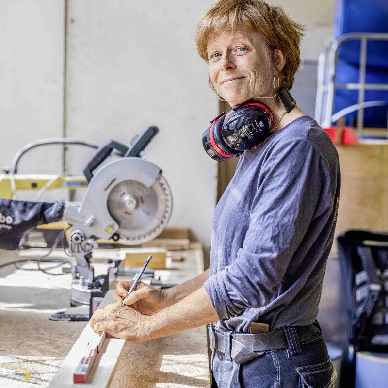 Frau steht in Werkstatt mit Geh&ouml;rschutz um den Hals und misst Holz aus