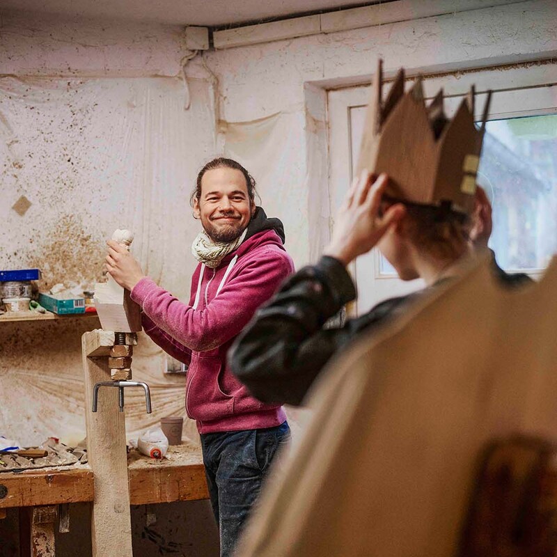 Mann in Werkstatt mit rotem Oberteil schaut Frau an, die eine Krone aus Pappe auf dem Kopf hat