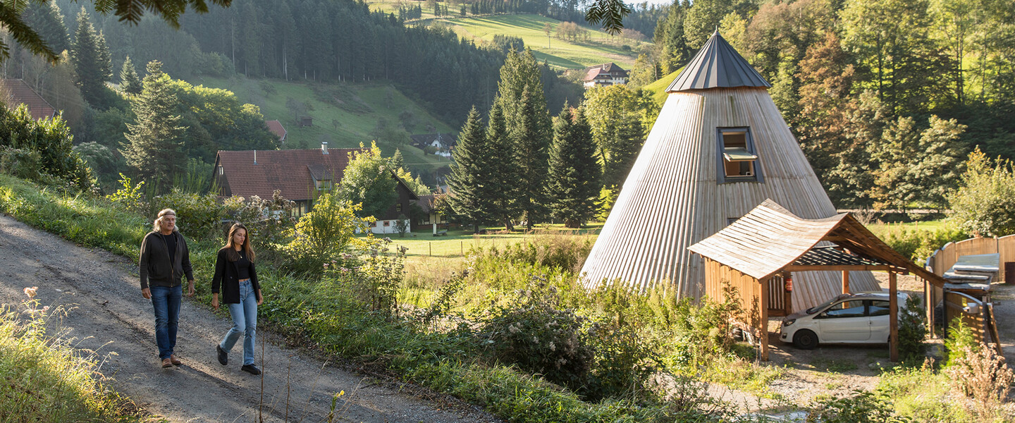 Mann und jüngere Frauen laufen einen Waldweg entlang, daneben steht ein Wohntipi aus Holz