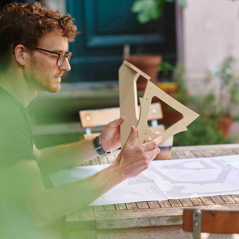 Mann mit Brille sitzt am Tisch und hält Holzteil in den Händen