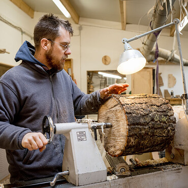 Mann mit Brille und Bart steht in einer Werkstatt und spannt einen Holzstamm in eine Drechselmaschine