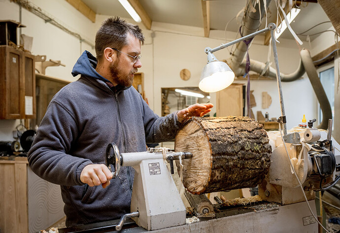 Mann mit Brille und Bart steht in einer Werkstatt und spannt einen Holzstamm in eine Drechselmaschine
