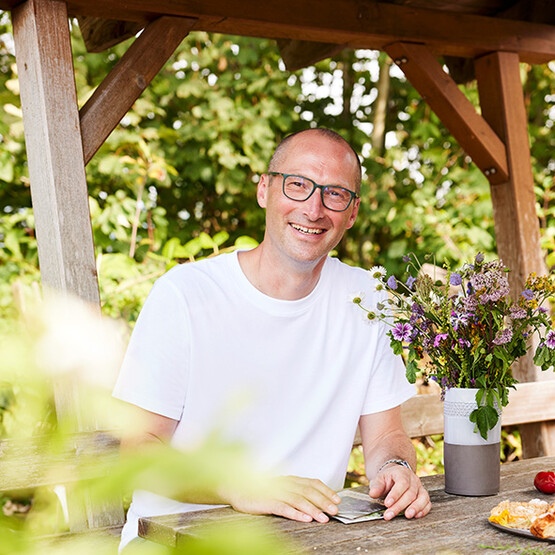 Mann mit wei&szlig;em T-Shirt und Brille (Johannes Huber von der der Firma Samen Maier) sitzt im Garten an einem Holztisch.