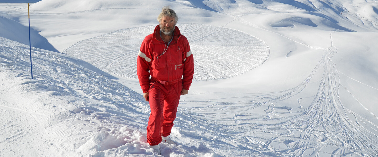 Älterer Mann mit Bart (Schneekünstler) und rotem Skianzug steht auf einem schneebedeckten Hügel in den Bergen