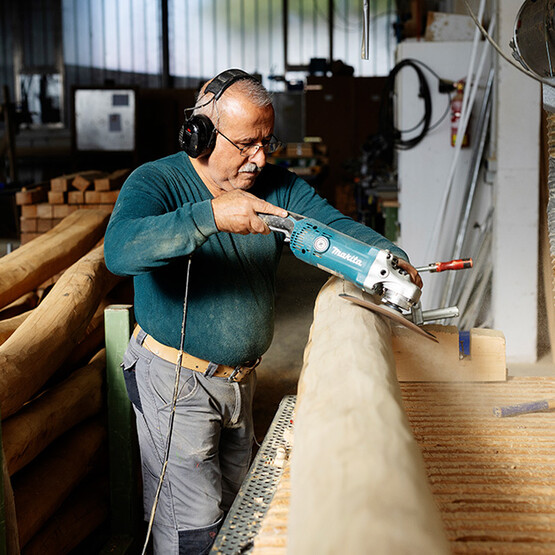 Älterer Mann mit Brille bearbeitet in einer Holzwerkstatt einen Holzstamm aus Robinie mit einem Schleifgerät