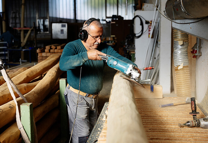 Älterer Mann mit Brille bearbeitet in einer Holzwerkstatt einen Holzstamm aus Robinie mit einem Schleifgerät