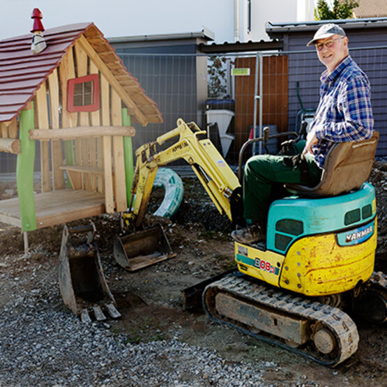 Älterer Mann (Dierk Mehrtens) sitzt auf einem Mini-Bagger auf einer Spielplatz-Baustelle