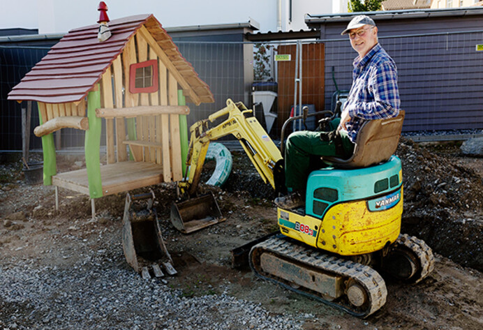 Älterer Mann (Dierk Mehrtens) sitzt auf einem Mini-Bagger auf einer Spielplatz-Baustelle