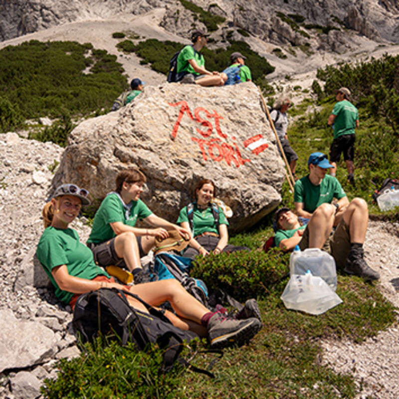 Sieben Freiwillige in grünen T-Shirts sitzen während einer Arbeitspause vor einem großen Stein im Hochgebirge