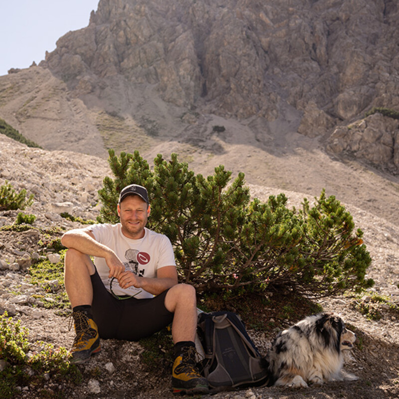 Ein Mann sitzt vor einem Busch im Hochgebirge, daneben ein Hund