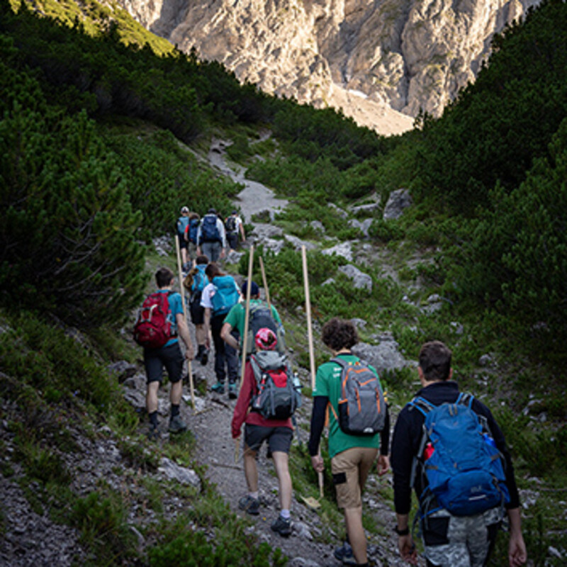 Wanderer mit Rucksäcken und Werkzeugen wandern einen Schotterweg im Hochgebirge hinauf