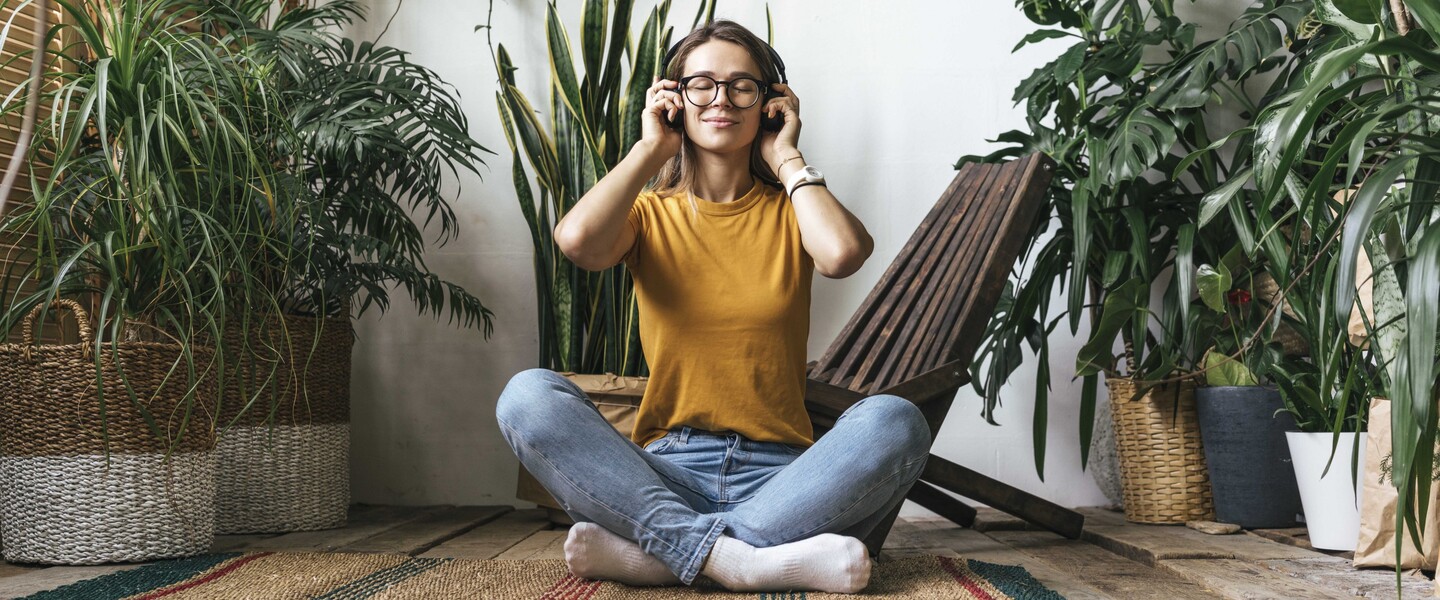 Frau sitzt zwischen Zimmerpflanzen auf Teppich und h&ouml;rt Musik